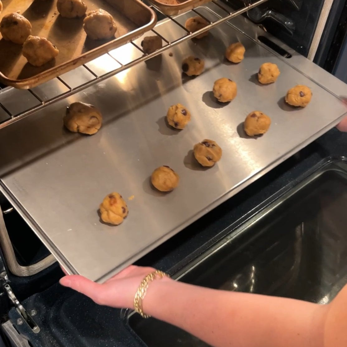 Person placing uncooked cookie dough balls on a titanium cutting board inside an oven.